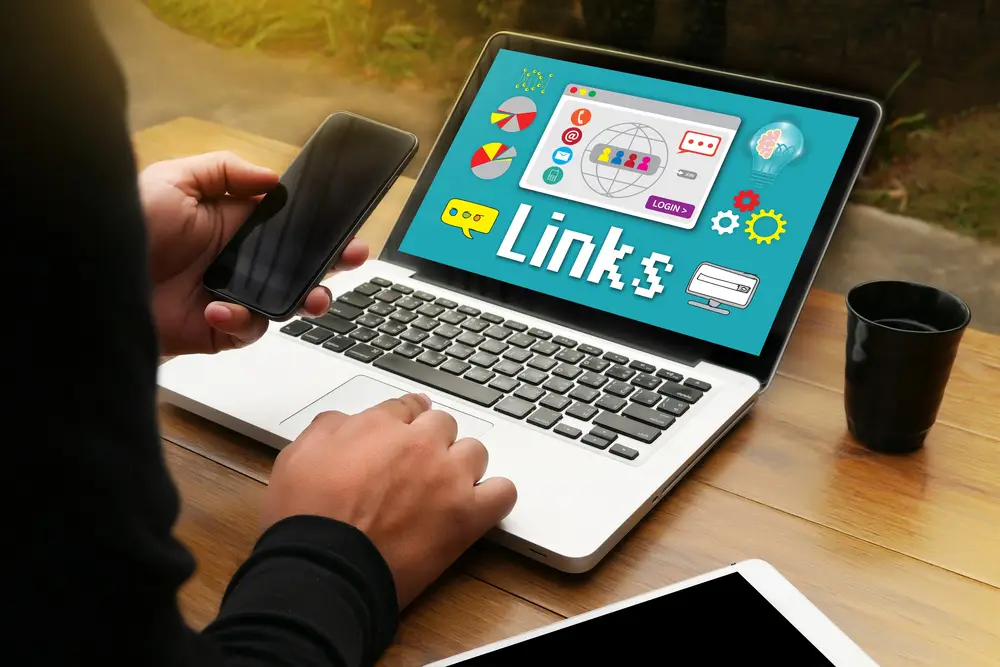 Persona che tiene uno smartphone nero in una mano mentre digita su un laptop con l'altra mano, su una scrivania di legno chiaro. Sullo schermo del laptop è visualizzata un'interfaccia colorata su sfondo turchese con la parola "Link" prominente al centro, circondata da varie icone digitali che rappresentano elementi di networking e comunicazione: grafici a torta, icone social, globo terrestre con figure umane stilizzate, simboli di chat, ingranaggi, e altre icone legate al marketing digitale e ai collegamenti web. Sulla scrivania si vedono anche una tazza nera e un tablet bianco. L'ambiente suggerisce un workspace dedicato alla gestione di link building o strategie SEO, con un'atmosfera professionale e moderna tipica del lavoro nel digital marketing.